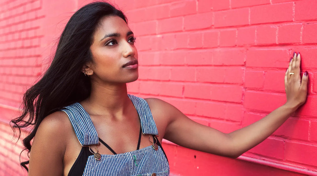 A woman with flowing dark hair and striped overalls leans one manicured hand on a red brick wall and stares up to the sky, dreaming that the future of women's fashion could be this comfortable.